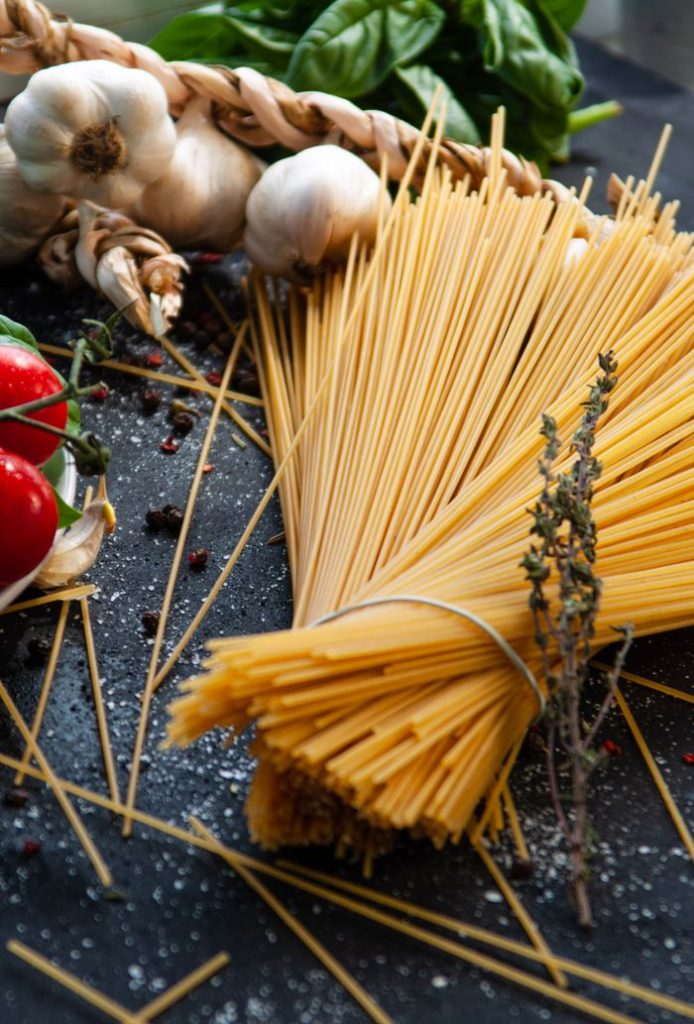 Bundle of raw spaghetti with tomatoes, garlic, and herbs on a black surface.